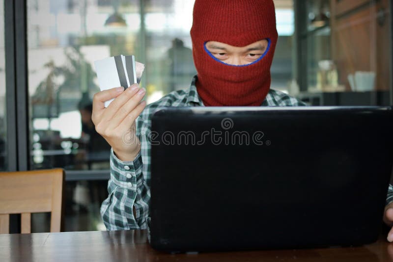 Selective focus on hands of Masked hacker wearing a balaclava holding credit card between stealing data from laptop. Internet crim royalty free stock photo
