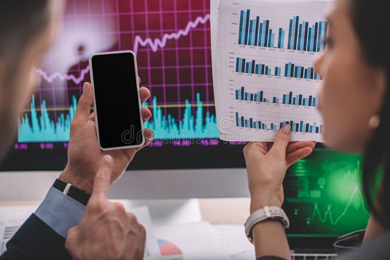 Focus of information security analyst pointing at smartphone near colleague with charts on paper at table stock photo