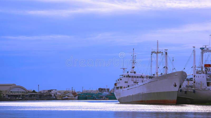 Large fishing boats with many vessel moored at harbor in industrial community along the coast against blue sky in evening time