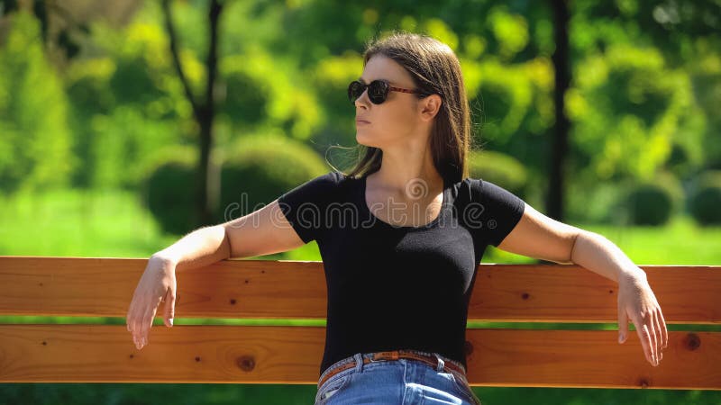 Self-confident woman in dark sunglasses sitting on bench in park looking around