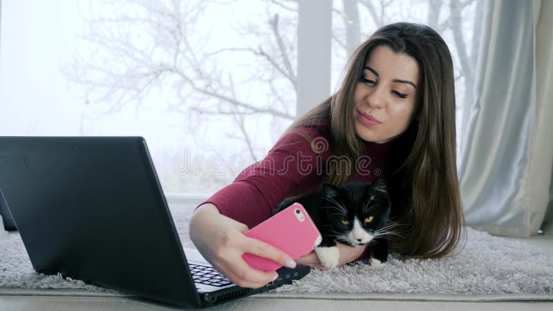 Selfie with cat, long-haired girl is photographed on smartphone with pet lying at floor on background of window