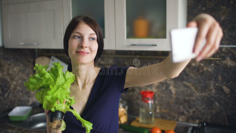 Selfie engraçado do tiro da dona de casa da mulher com salada verde ao cozinhar na cozinha em casa dentro