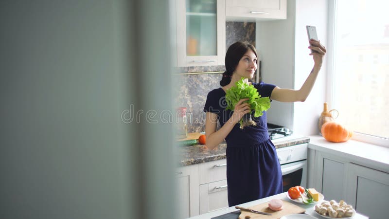 Selfie engraçado do tiro da dona de casa da mulher com salada verde ao cozinhar na cozinha em casa dentro