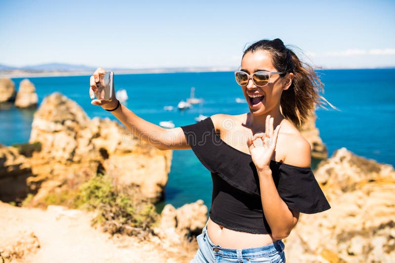Selfie latino della presa della donna di bellezza dal telefono alla cima di bella scogliera s ed alle rocce dell'Oceano Atlantico fotografie stock Selfie latino della presa della donna di bellezza dal telefono alla cima di bella scogliera s ed alle rocce dell'Oceano Atlantico fotografie stock