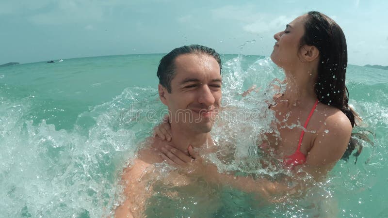 Selfie smiling man and woman swimming in big ocean waves while vacationing on island. Woman holds tightly to companion