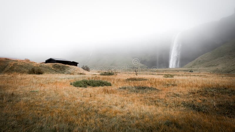 Seljalandsfoss Wasserfall an einem nebligen Tag. Seljalandsfoss scheint an einem nebligen Tag in Eisland von den Wolken zu fallen. Es gibt ein großes Grasfeld im Vordergrund. ein Bauernhaus aus dem Feld im Hintergrund erhebt. es fühlt sich melancholisch an.
