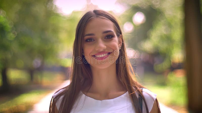 A senhora agradável está estando no parque no dia, olhando na câmera, sorriso, backgrouns borrados