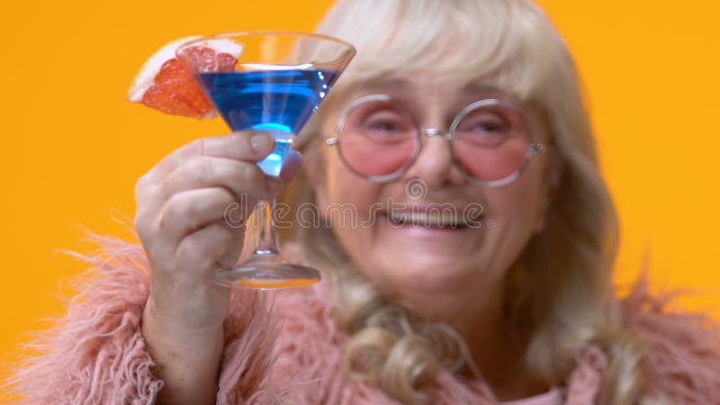 A senhora envelhecida chique que mostra o vidro de cocktail azul à disposição, avó à moda relaxa