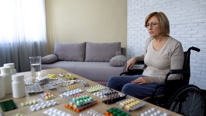 Senhora idosa só que senta-se na cadeira de rodas perto da tabela com a medicina que sente comprimida fotos de stock Senhora idosa só que senta-se na cadeira de rodas perto da tabela com a medicina que sente comprimida fotos de stock