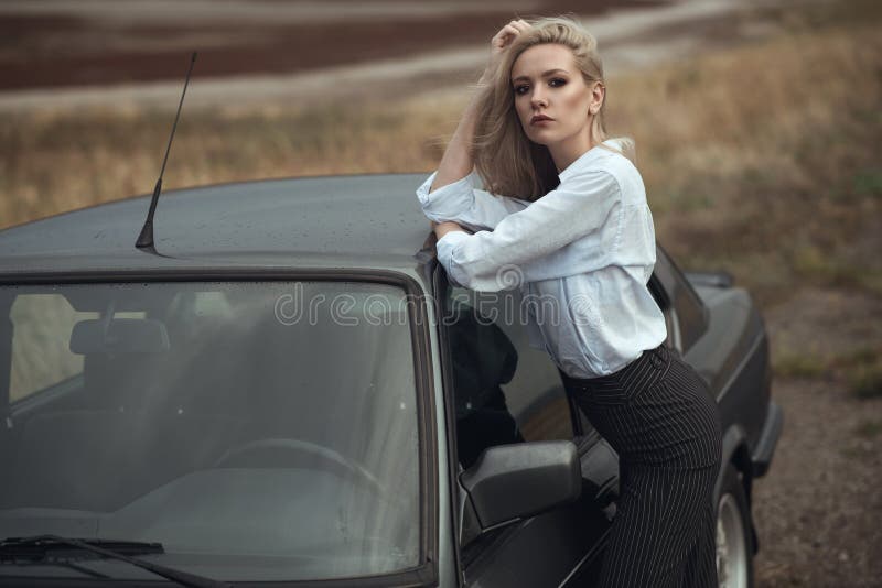 Senhora loura bonita em calças waisted altas listradas pretas e na blusa branca que inclinam-se na parte superior de seu carro ve imagem de stock Senhora loura bonita em calças waisted altas listradas pretas e na blusa branca que inclinam-se na parte superior de seu carro ve imagem de stock