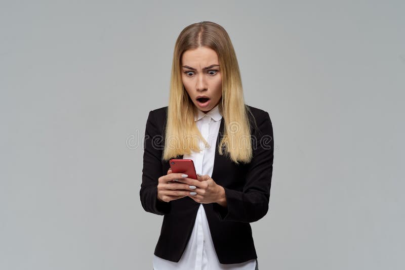 A senhora loura de cabelos compridos do negócio no vestido formal e na blusa branca que guardam um smartphone e é chocada para le imagens de stock A senhora loura de cabelos compridos do negócio no vestido formal e na blusa branca que guardam um smartphone e é chocada para le imagens de stock