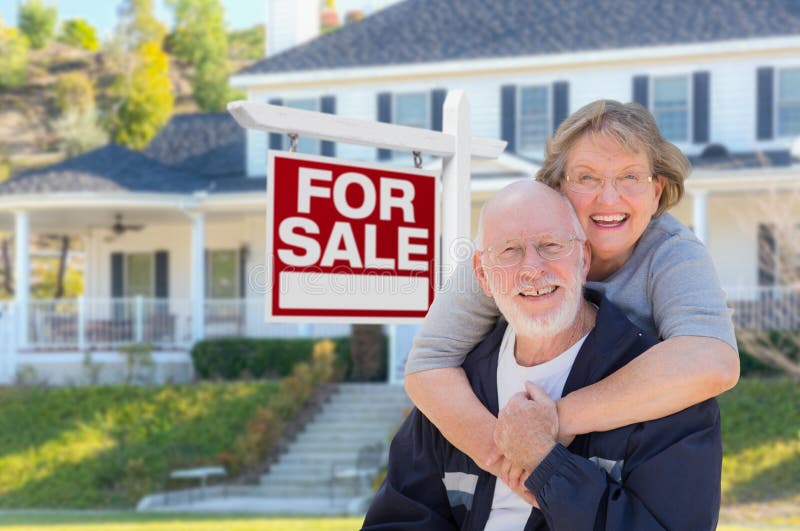 Senior Adult Couple in Front of Home For Sale Real Estate Sign and Beautiful House. Senior Adult Couple in Front of Home For Sale Real Estate Sign and Beautiful House.