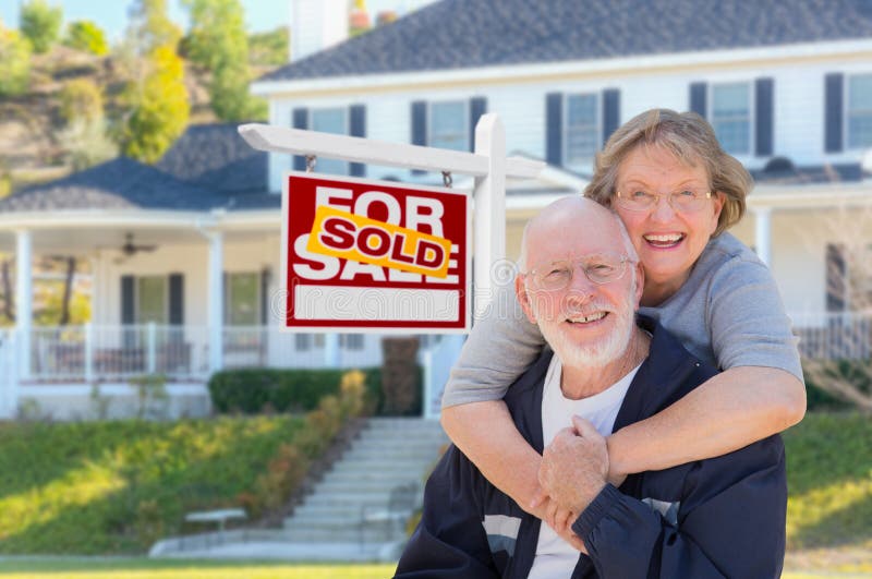 Senior Adult Couple in Front of Sold Home For Sale Real Estate Sign and Beautiful House. Senior Adult Couple in Front of Sold Home For Sale Real Estate Sign and Beautiful House.