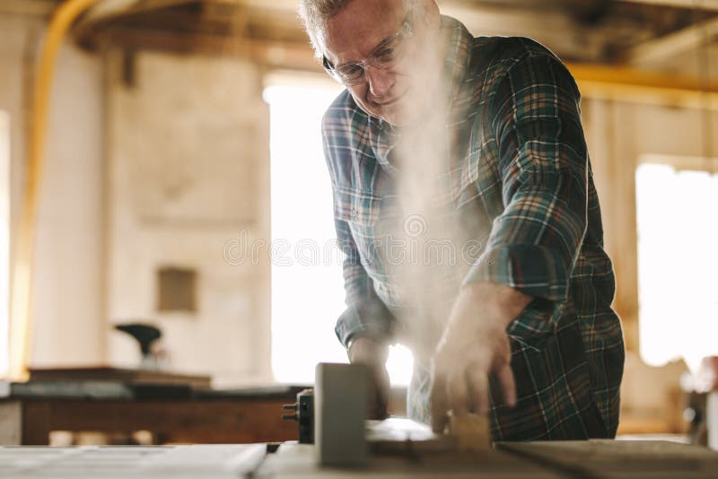 Mature male carpenter working on table saw machine in carpentry workshop. Senior man cutting wood on table saw machine. Mature male carpenter working on table saw machine in carpentry workshop. Senior man cutting wood on table saw machine