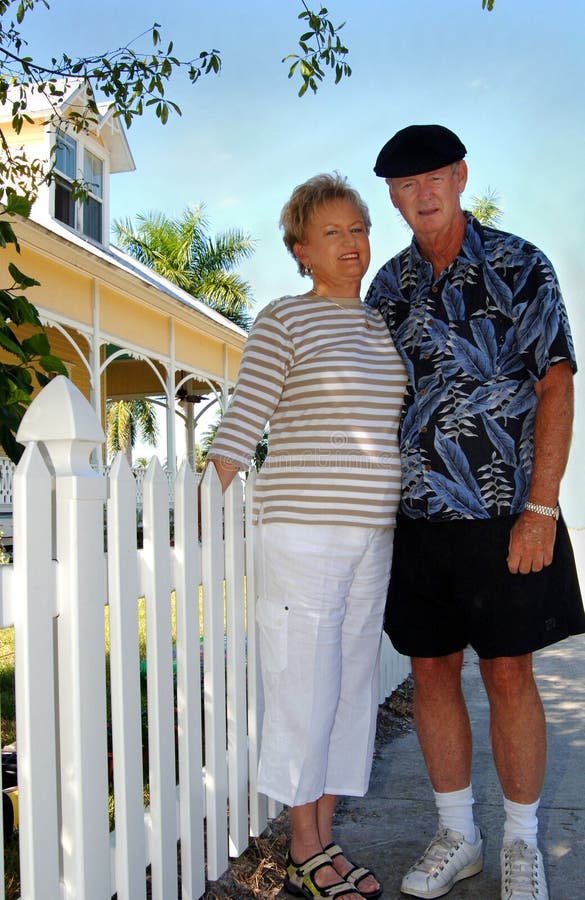 A happy senior couple standing in front of white picket fence and house. A happy senior couple standing in front of white picket fence and house.