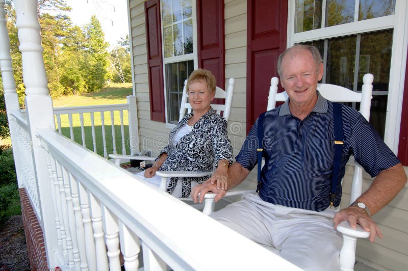 A happy senior couple holding hands and sitting on the front porch of a home in rocking chairs. A happy senior couple holding hands and sitting on the front porch of a home in rocking chairs