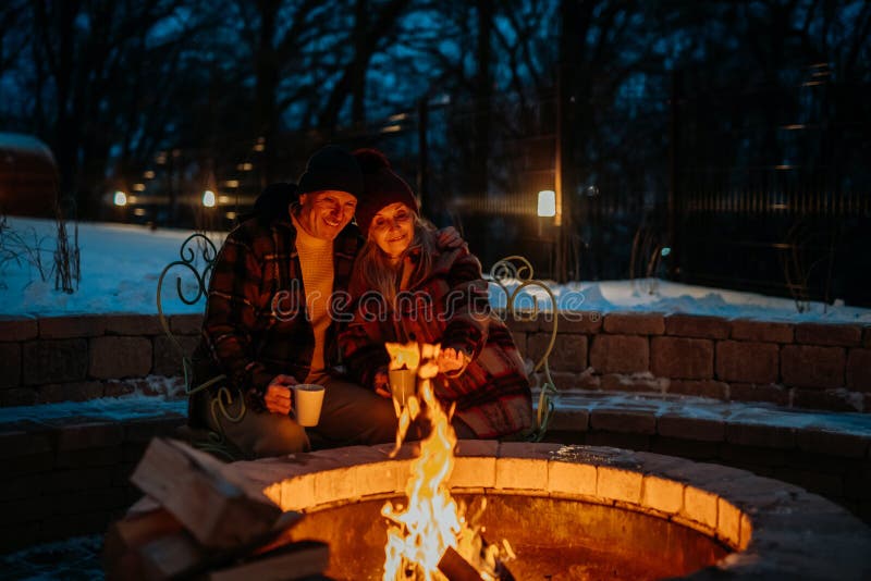 Senior couple sitting and heating together at outdoor fireplace in winter evening.