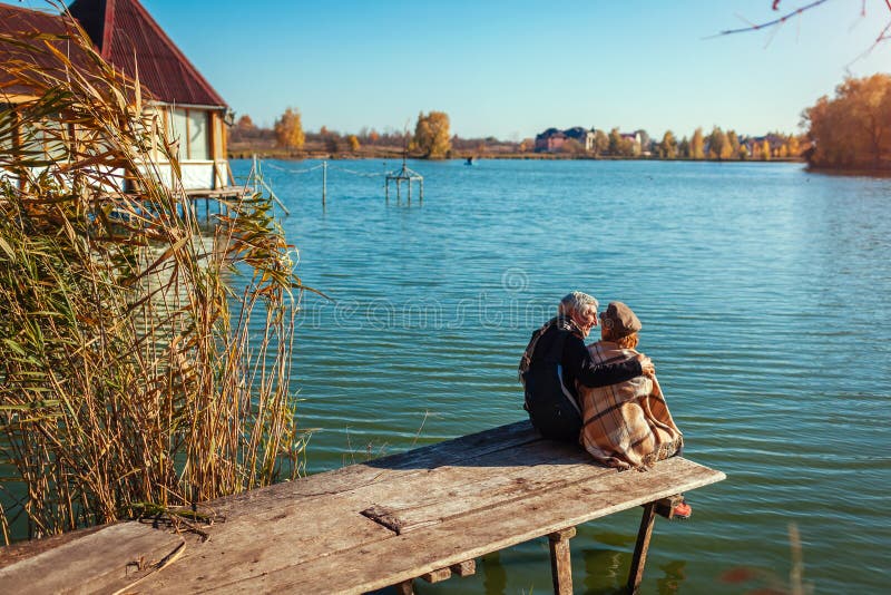 Senior family couple relaxing by autumn lake. Happy man and woman enjoying nature and hugging sitting on pier