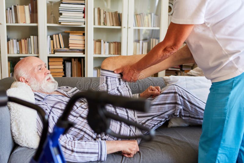 Senior in pajamas on the sofa receives physiotherapy for knee osteoarthritis from nurse. Senior in pajamas on the sofa receives physiotherapy for knee osteoarthritis from nurse