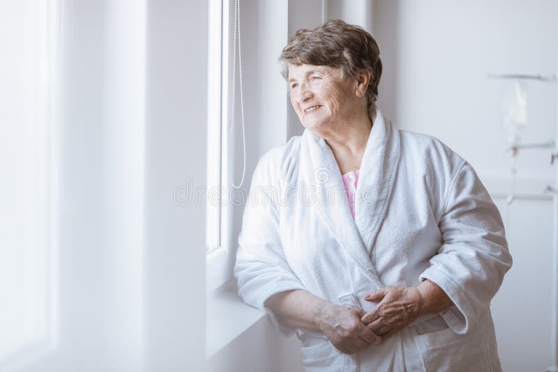 Senior grey lady wearing white bathrobe standing by the window at nursing home stock photo Senior grey lady wearing white bathrobe standing by the window at nursing home stock photo