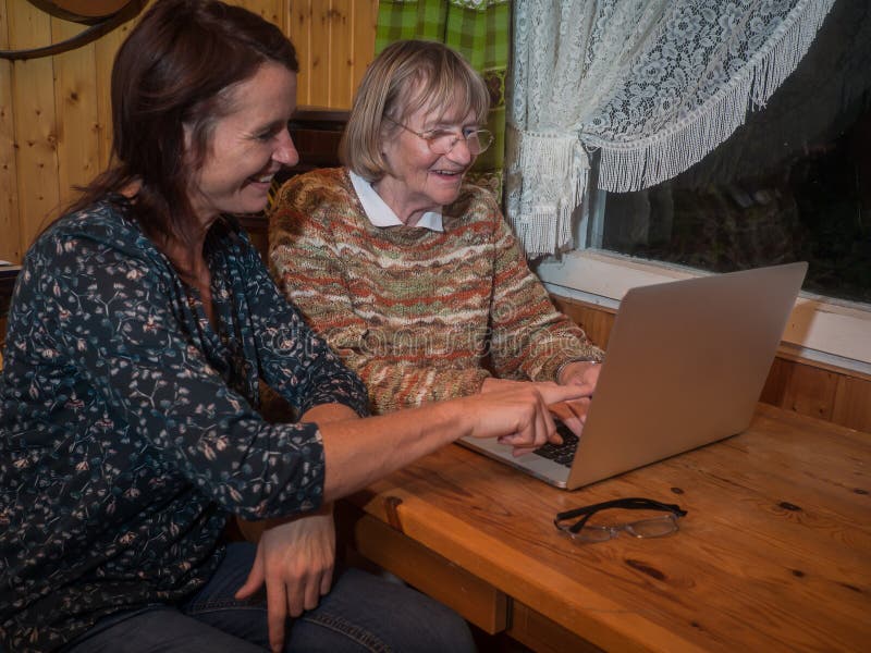 Senior and mature women having fun using a laptop. They are sitting on a wooden table in a small living room in the evening. Senior and mature women having fun using a laptop. They are sitting on a wooden table in a small living room in the evening.