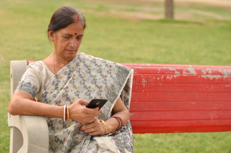 Senior Indian Bengali woman working on a mobile phone, experimenting with technology on a red park bench in an outdoor setting