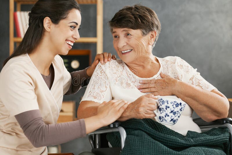 Senior lady in wheelchair holding cup of herbal tea and smiling to pretty volunteer at nursing home royalty free stock photo