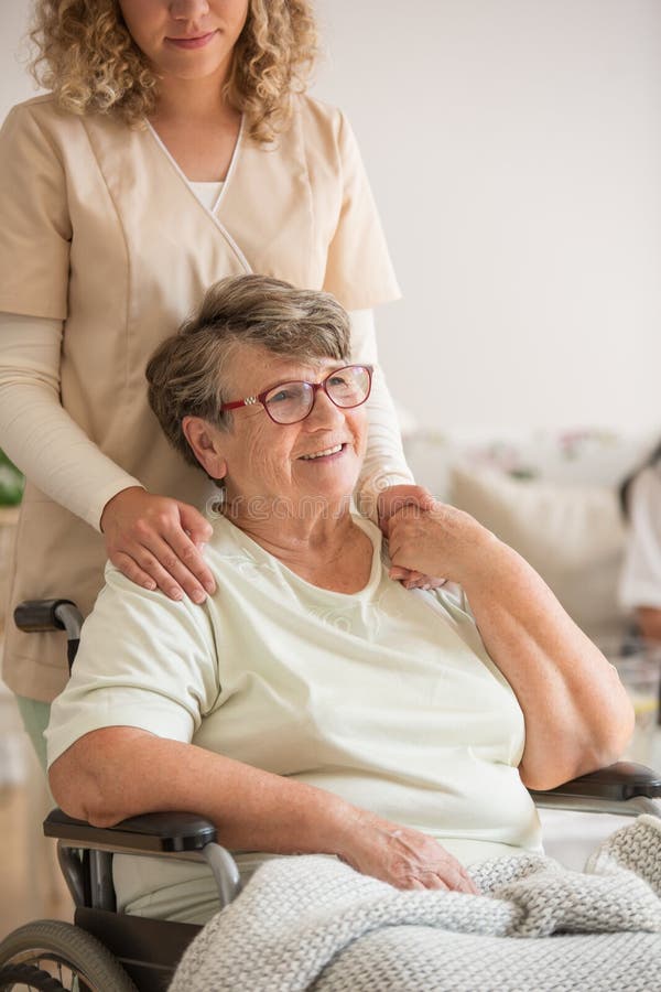 Senior lady on wheelchair with young volunteer in beige uniform supporting her royalty free stock photos