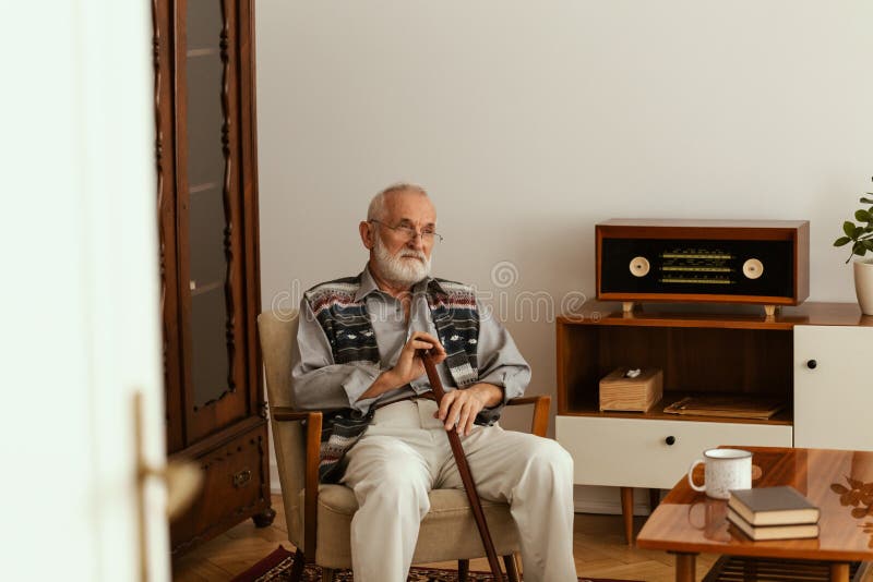 Senior man with grey hair and beard and with walking stick sitting on armchair alone in his apartment royalty free stock images Senior man with grey hair and beard and with walking stick sitting on armchair alone in his apartment royalty free stock images