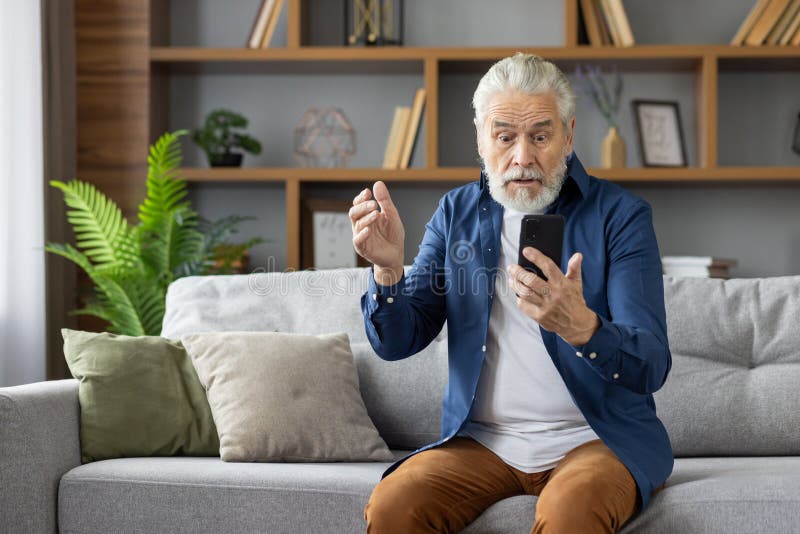 Elderly man with grey hair sits on a sofa, looking shocked and confused as he reads a distressing message on his smartphone in a modern living room. Elderly man with grey hair sits on a sofa, looking shocked and confused as he reads a distressing message on his smartphone in a modern living room.