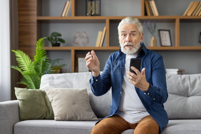 Elderly Caucasian man looking puzzled and stressed while looking at his mobile phone, sitting on a sofa in a well-lit living room. Elderly Caucasian man looking puzzled and stressed while looking at his mobile phone, sitting on a sofa in a well-lit living room.