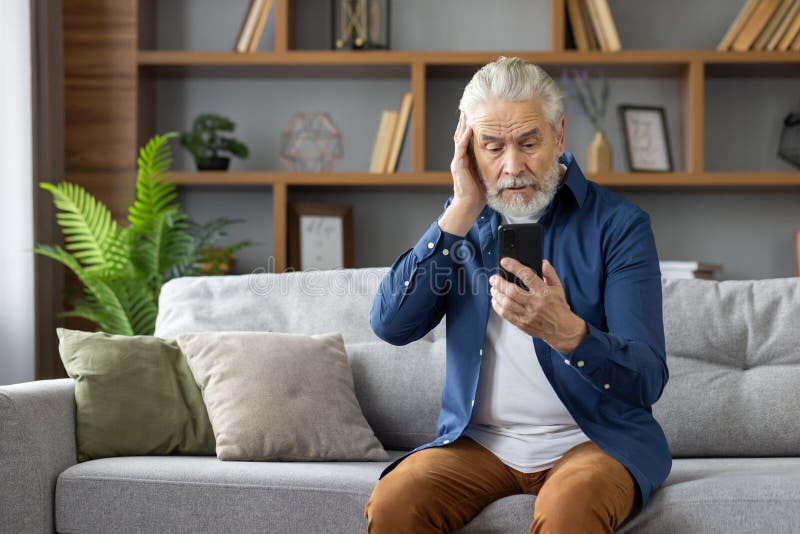 An elderly man sits on a sofa, looking confused and worried as he reads a message on his mobile phone, portraying a moment of concern. An elderly man sits on a sofa, looking confused and worried as he reads a message on his mobile phone, portraying a moment of concern.