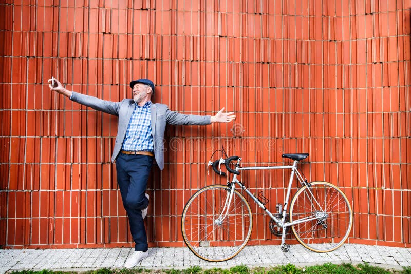 Senior man with smartphone and bicycle against brick wall. Sneakers, confident.