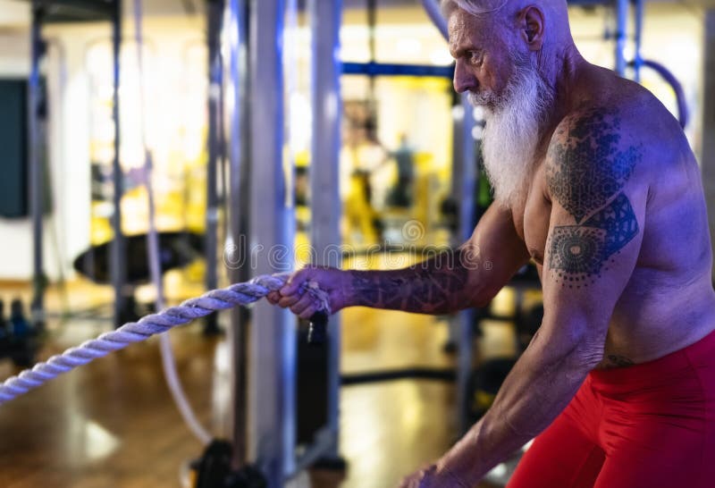 Senior man training with battle rope exercise inside gym - Fit mature male workout in wellness club center