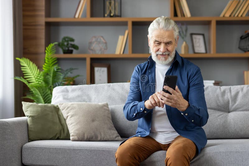 A cheerful elderly Caucasian man sits comfortably on a grey sofa using a smartphone in a well-decorated living room with green plants. A cheerful elderly Caucasian man sits comfortably on a grey sofa using a smartphone in a well-decorated living room with green plants.