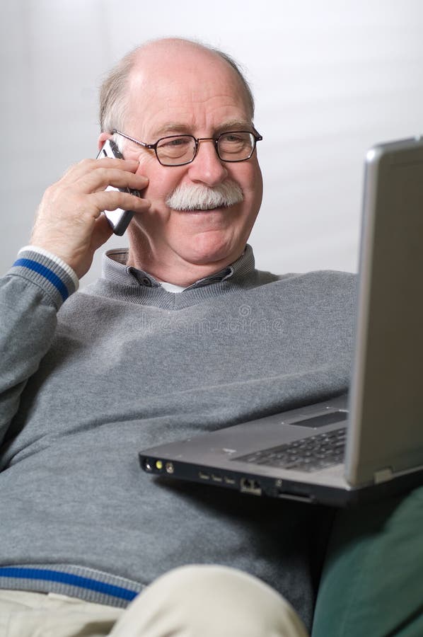 Senior man working on laptop and calling by phone while sitting on the couch