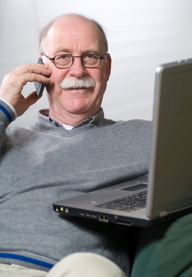 Senior man working on laptop and calling by phone while sitting on the couch