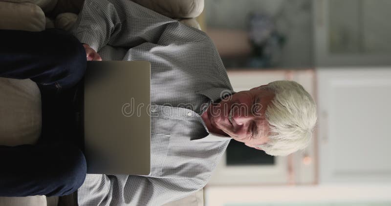 Senior Mann mit vertikaler Sicht sitzt auf dem Sofa und tippt auf dem Laptop