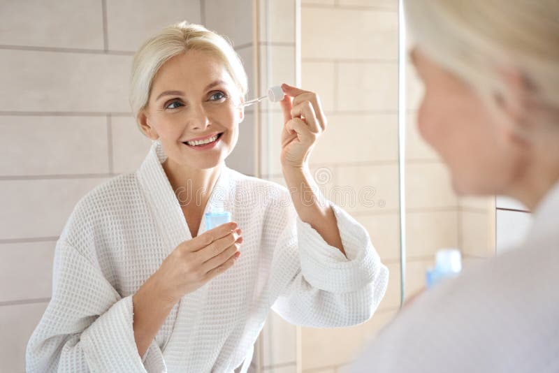 Mature senior European woman with pipette serum essence looking at mirror.