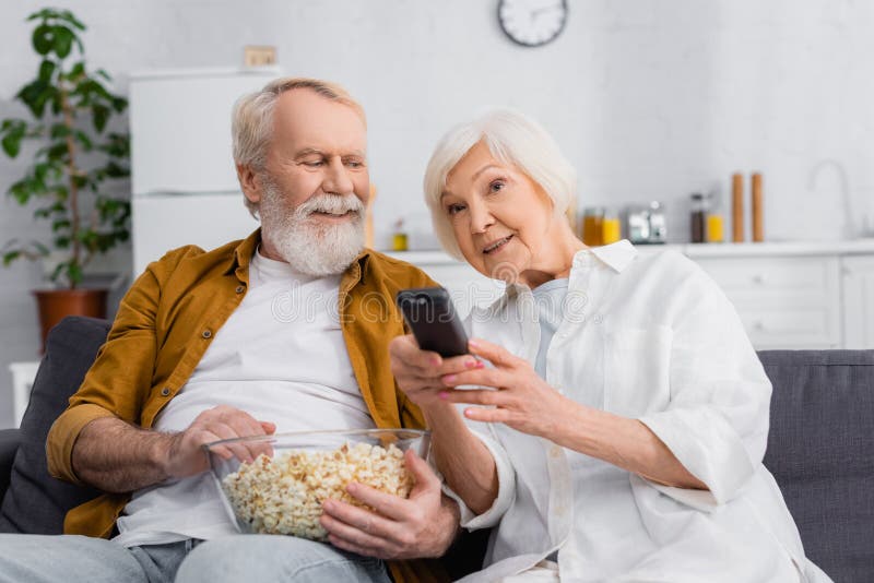 Senior woman clicking channels near husband with bowl of popcorn on couch.