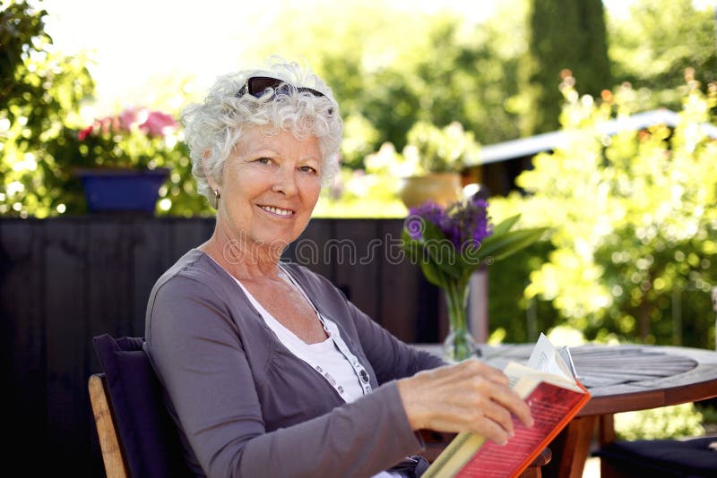 Senior woman sitting on a chair in backyard garden holding a book and looking at camera smiling. Senior woman sitting on a chair in backyard garden holding a book and looking at camera smiling