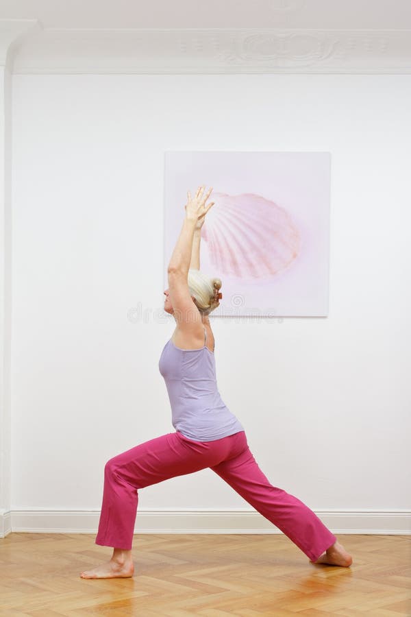 Senior woman with gray hair doing yoga exercises at home in front of a wall with a picture, asana warrior I. Senior woman with gray hair doing yoga exercises at home in front of a wall with a picture, asana warrior I