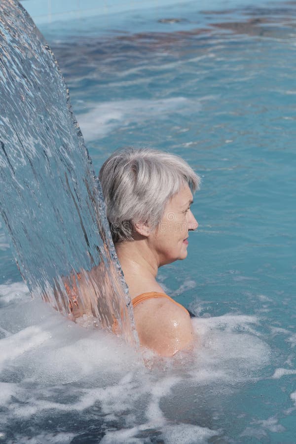 Senior woman with gray hair near falling flow of water in outdoor thermal swimming pool.