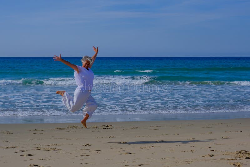 Senior woman with greay hair and a white dress jumping in the air at the beach
