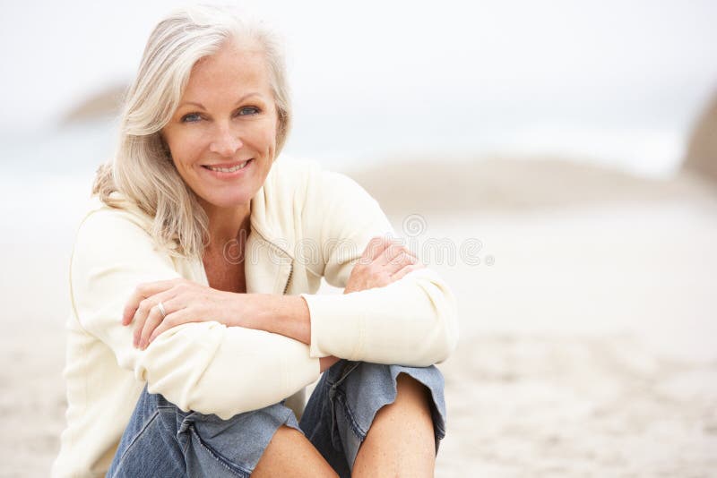 Senior Donna In Vacanza Seduto Sulla Spiaggia D'Inverno, E Sorridendo Alla Telecamera.