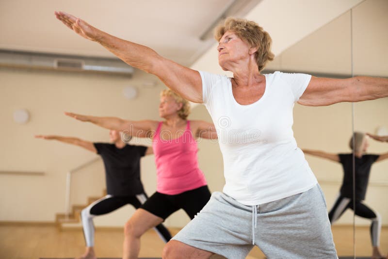 Concentrated senior woman performing standing lunging asana Virabhadrasana during yoga class with female group in fitness studio. Concentrated senior woman performing standing lunging asana Virabhadrasana during yoga class with female group in fitness studio.