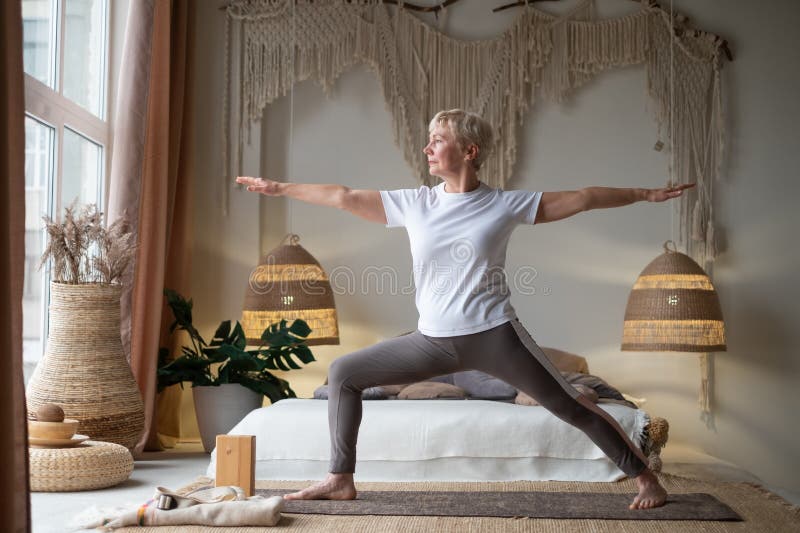 Senior woman practicing yoga, standing in Warrior two exercise, Virabhadrasana II pose, working out at home