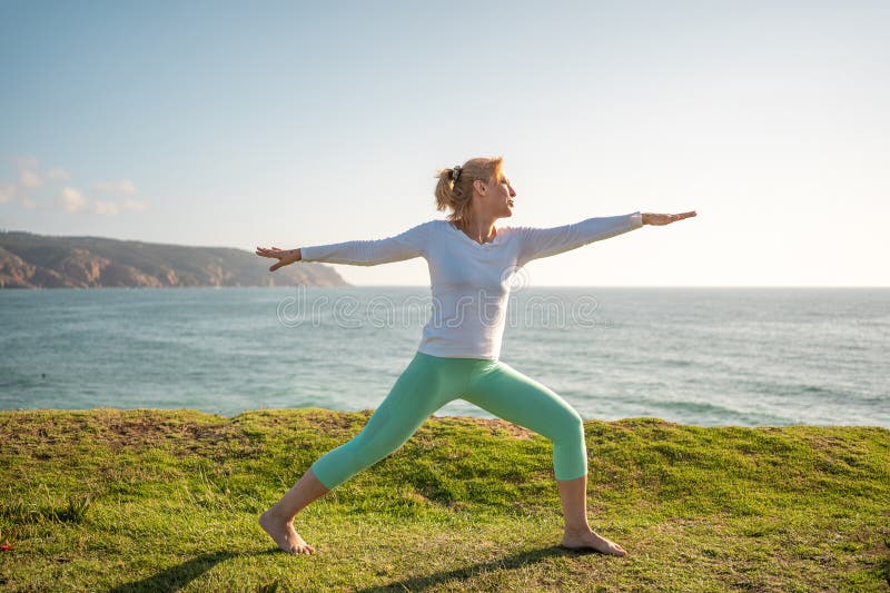 Senior woman pensioner practicing yoga warrior pose on beach. Performing virabhadrasana enjoying ocean view. Yogi female taking good care of her body and living healthy lifestyle. Senior woman pensioner practicing yoga warrior pose on beach. Performing virabhadrasana enjoying ocean view. Yogi female taking good care of her body and living healthy lifestyle.