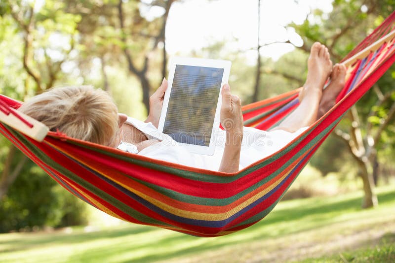 Senior Woman Relaxing In Hammock With E-Book During Summer