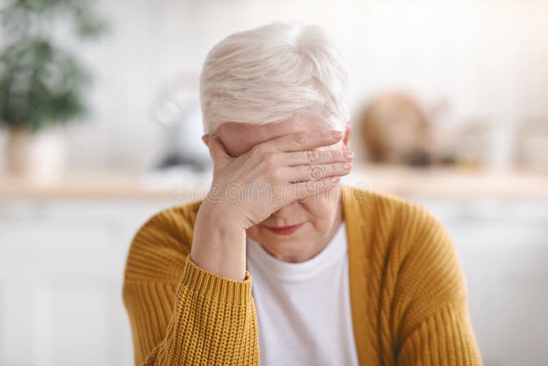 Senior grey-haired woman suffering from migraine, touching her forehead, kitchen interior, closeup. Old lady having headache, experiencing problems with health during climax, climacteric syndrome. Senior grey-haired woman suffering from migraine, touching her forehead, kitchen interior, closeup. Old lady having headache, experiencing problems with health during climax, climacteric syndrome